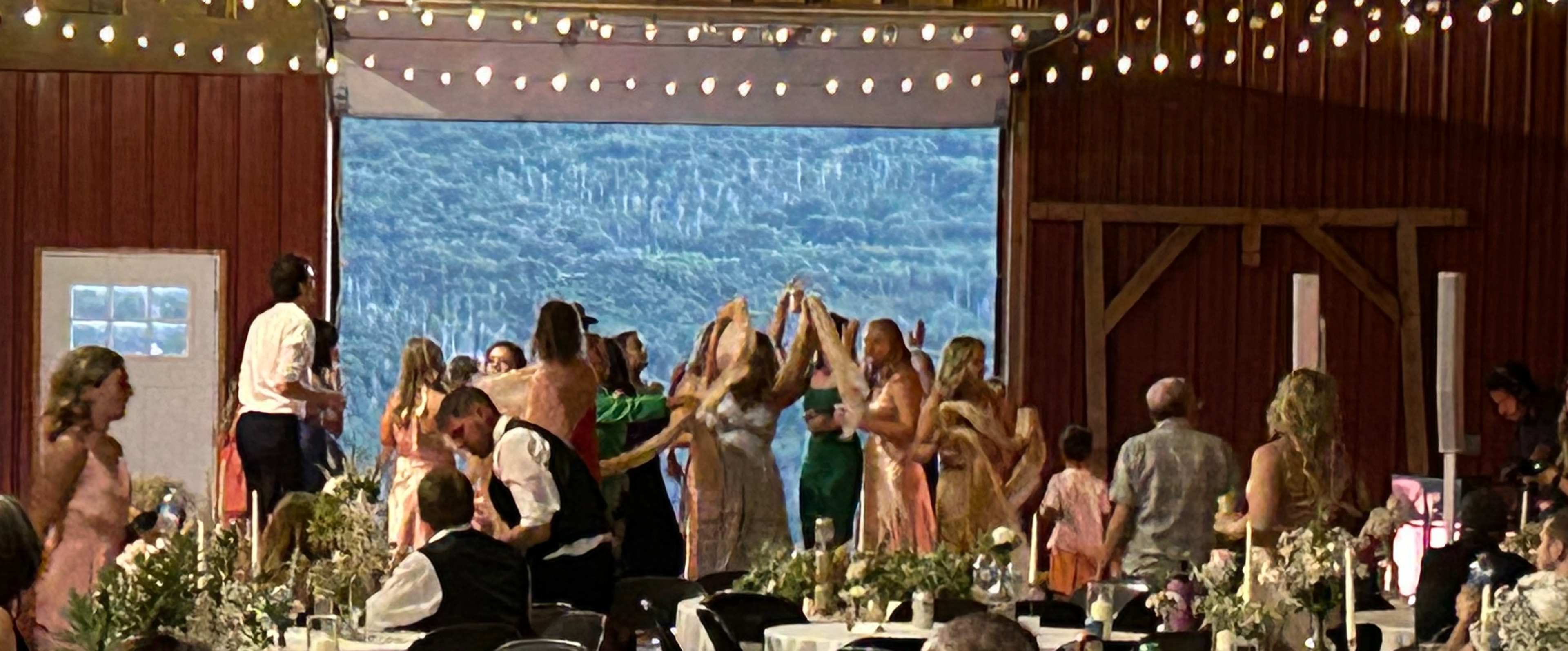 Guests dancing and celebrating inside the barn at the reception, string lights glowing overhead with the mountain forest visible through the open doors