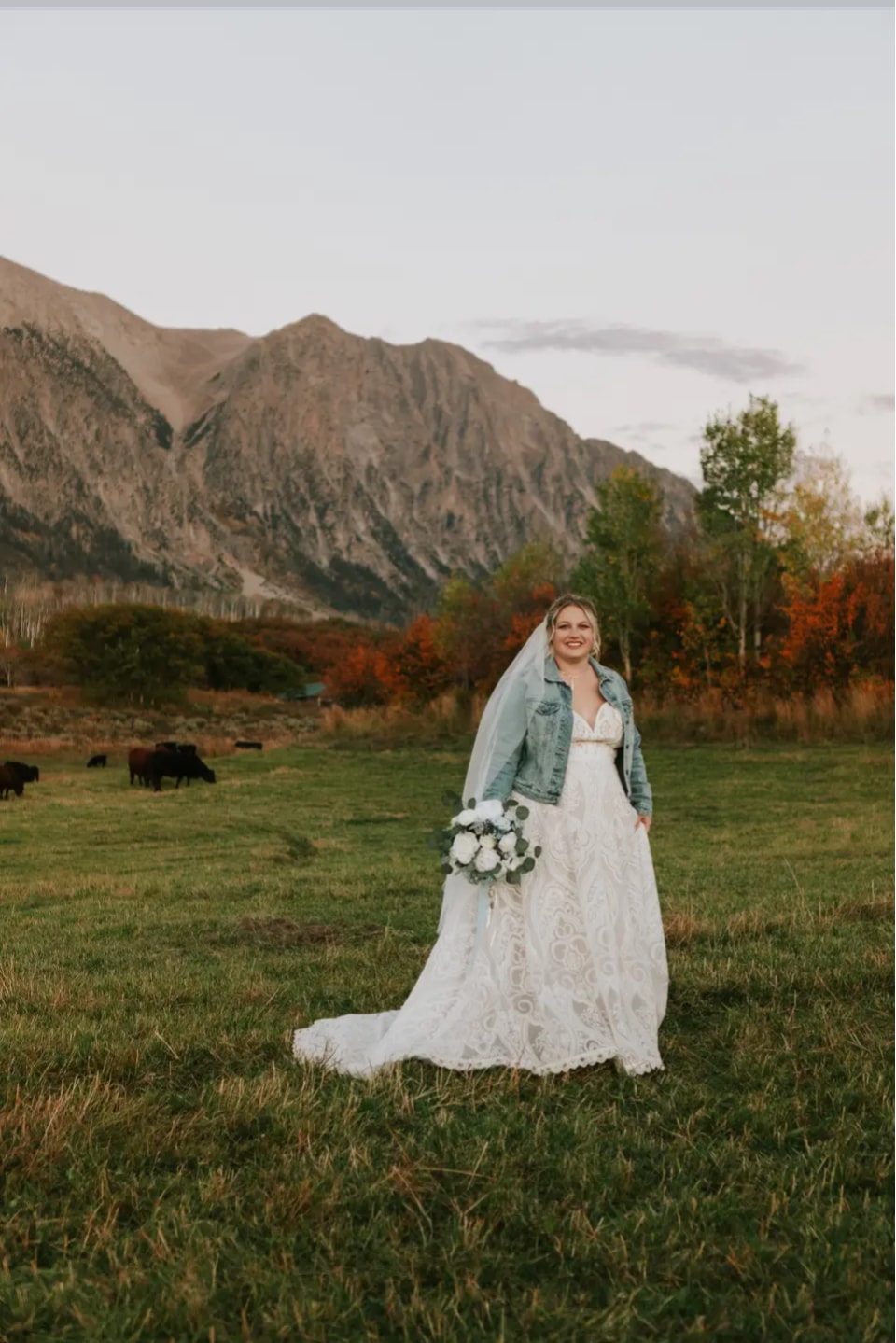 Bride in lace gown and denim jacket holding a white bouquet in the ranch pasture with Ragged Mountain at golden hour