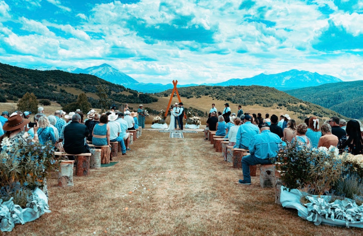 Wide shot of the outdoor ceremony with guests seated on log benches, couple at the wooden arch altar with Ragged Mountain peaks filling the horizon