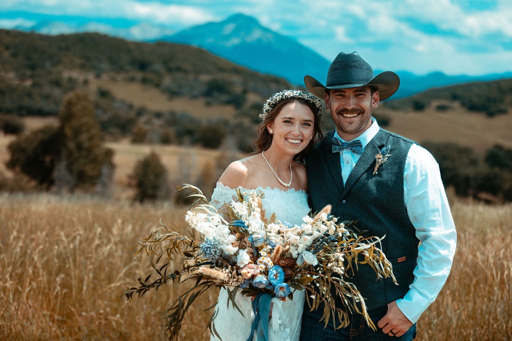 Bride and groom smiling together in a golden field with Ragged Mountain and blue sky behind them, bride holding a large wildflower bouquet