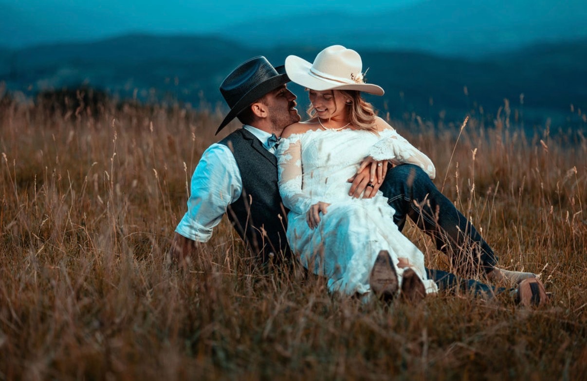 Couple sitting together in tall grass at dusk, bride in white dress and groom in cowboy hat, sharing a quiet moment