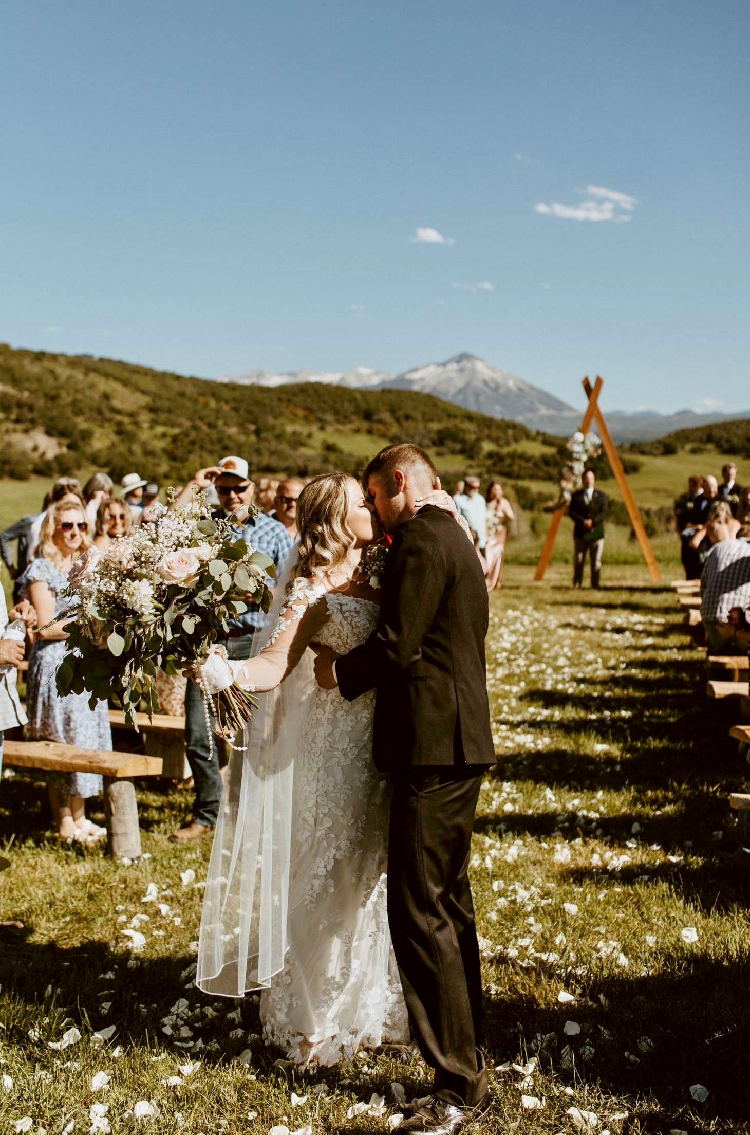 Bride and groom sharing their first kiss at the end of the petal-strewn aisle with Ragged Mountain and the wooden arch behind them
