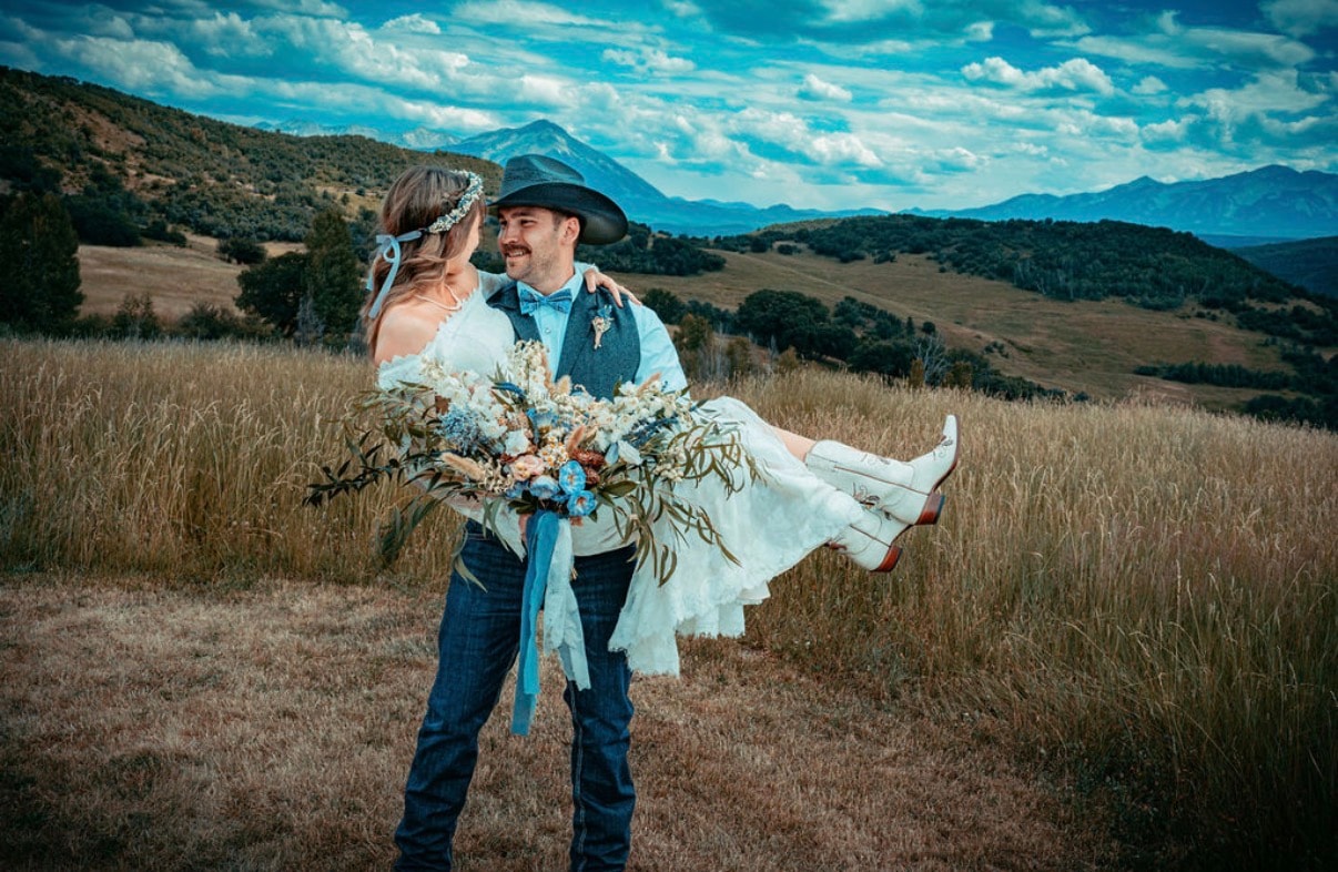 Groom carrying bride in his arms through a golden field with sweeping mountain valley views behind them