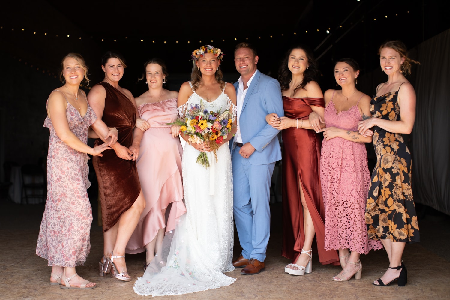 Bride with flower crown and colorful bouquet linked arm-in-arm with the wedding party inside the barn under string lights
